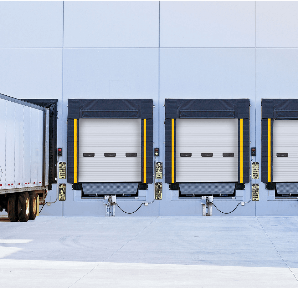 A white semi-trailer truck is backed up to one of several loading dock doors on a modern warehouse building, ready for loading or unloading.
